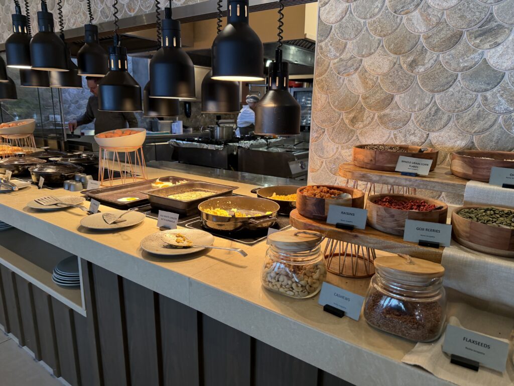 Buffet du petit-déjeuner avec fruits secs, graines, plats chauds et cuisiniers en arrière-plan.