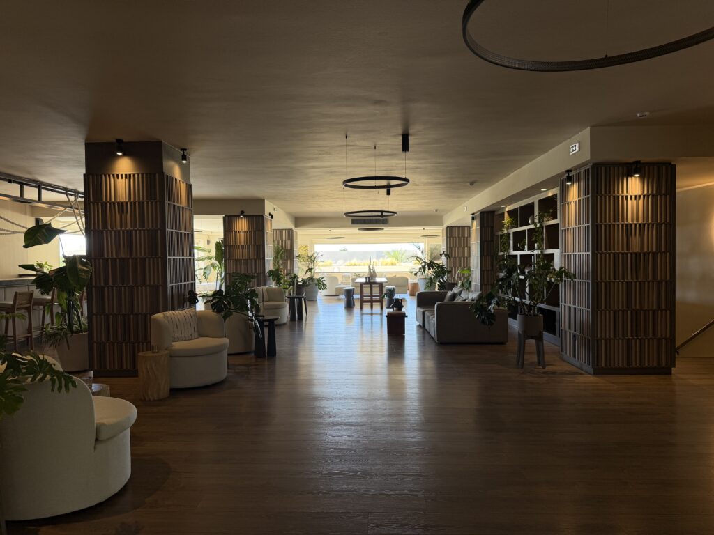 Salon lumineux du Numo Ierapetra avec mobilier beige, plantes vertes et ouverture sur la mer Égée.