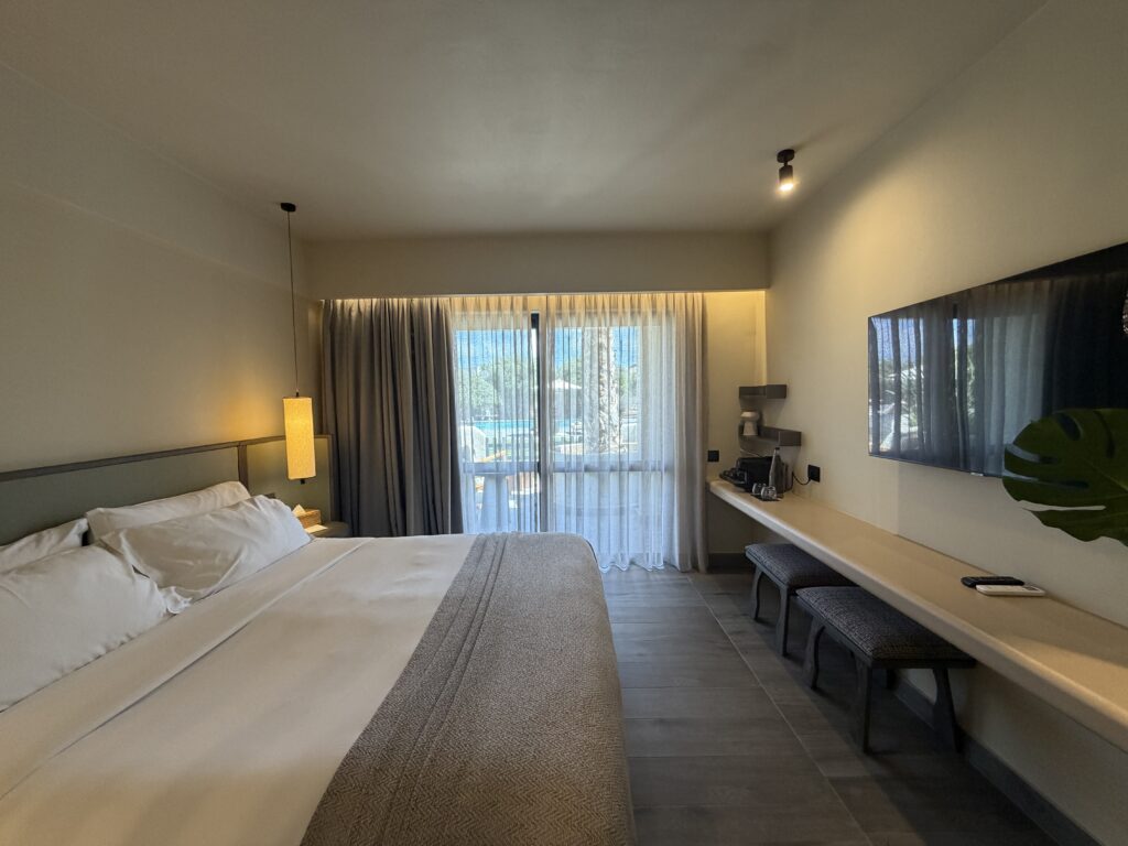 Chambre lumineuse du Numo Ierapetra avec grand lit, télévision murale et balcon donnant sur les palmiers.