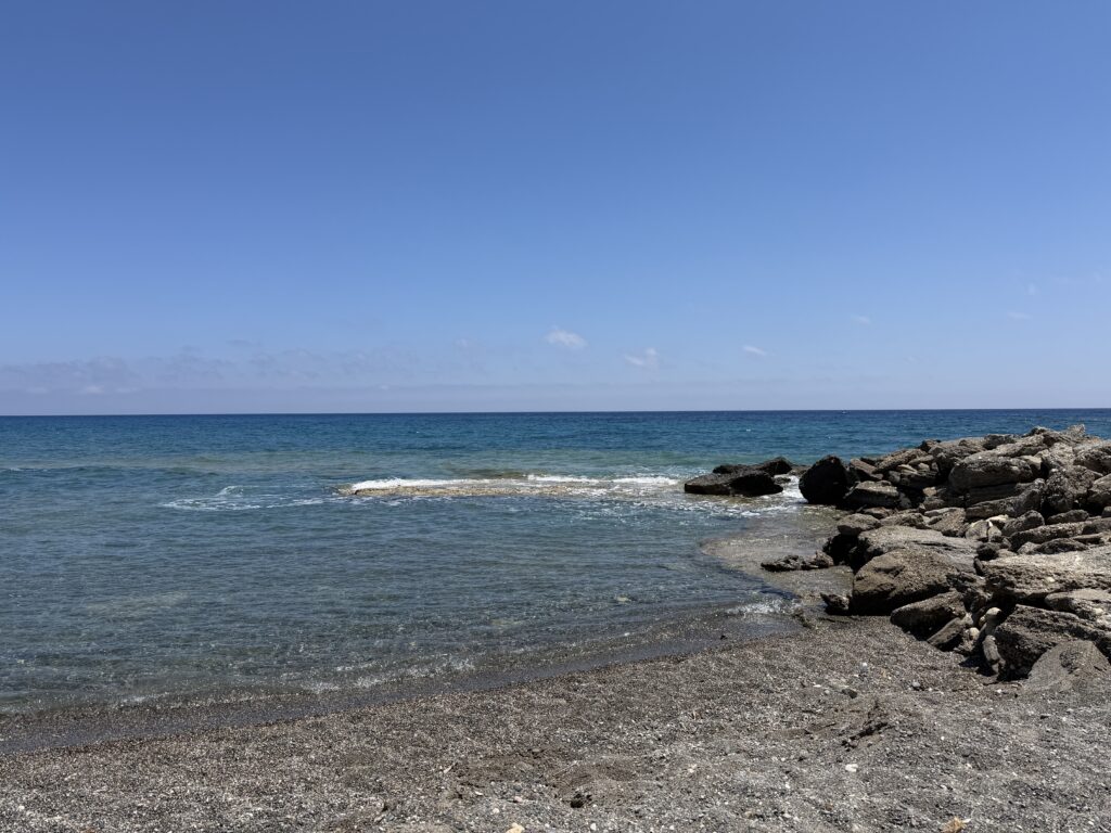 Plage de galets sombres bordée d’eau turquoise et rochers sous un ciel bleu clair.