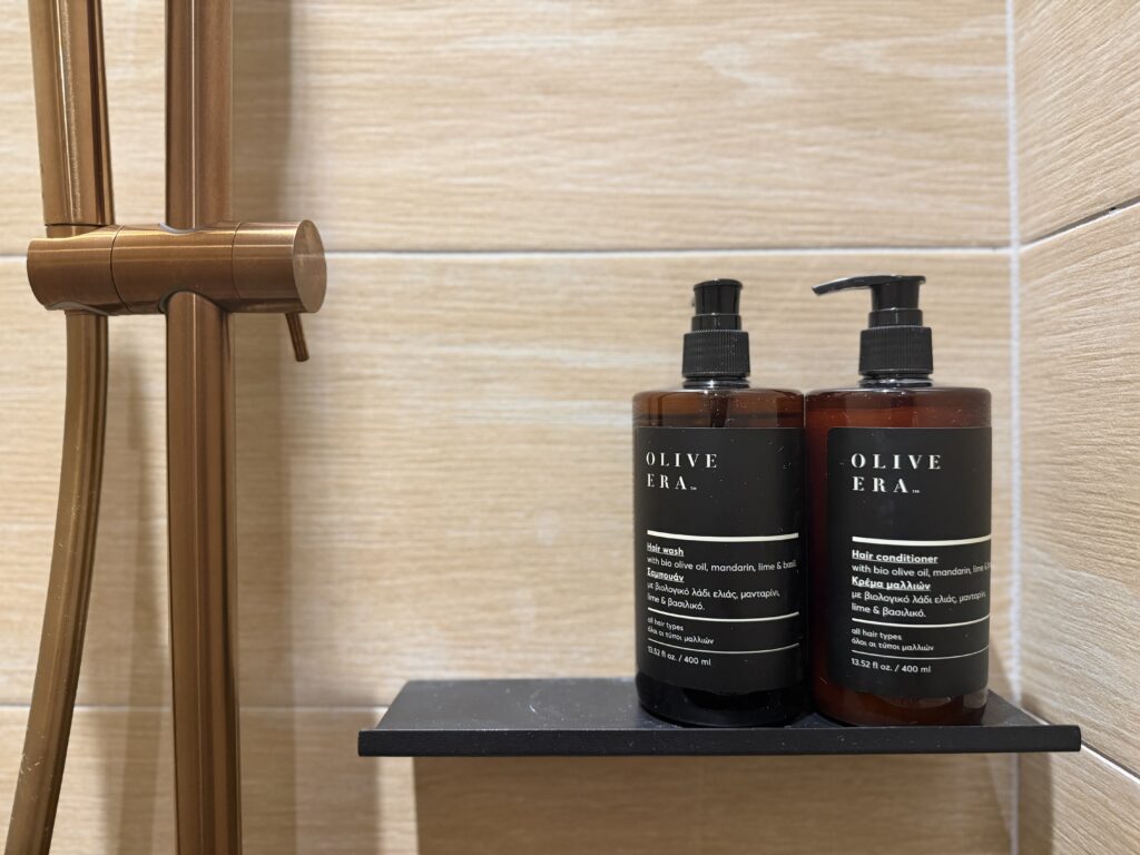 Deux flacons Olive Era sur une étagère noire dans une douche aux parois beige clair.