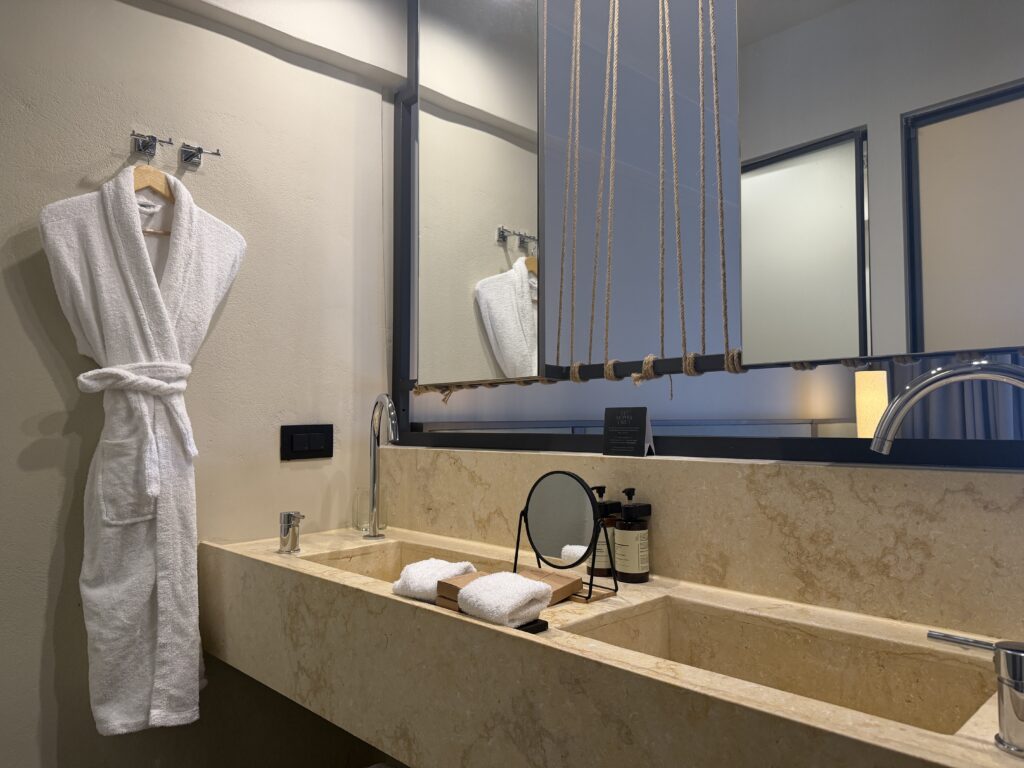 Salle de bain moderne en pierre beige avec grand miroir suspendu par des cordes naturelles.