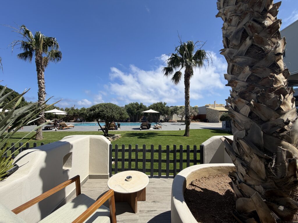 Terrasse privée avec fauteuils donnant sur pelouse, piscine et palmiers sous ciel bleu limpide.