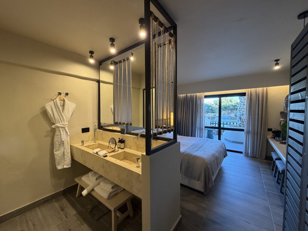 Chambre moderne du Numo Ierapetra avec lavabo en pierre, peignoir blanc et terrasse ensoleillée.