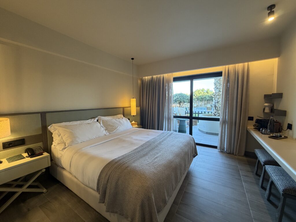 Chambre moderne avec grand lit double, éclairage doux, terrasse extérieure et vue sur palmiers crétois.