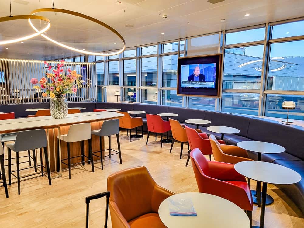 Espace détente du salon avec banquette, fauteuils orange et rouges et écran allumé